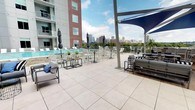 This modern urban residence offers a well-designed single-floor layout that balances contemporary style with functional living. The living area features warm hardwood flooring, large windows that invite abundant natural light, and a ceiling fan overhead. A tray ceiling detail adds architectural interest, while the open layout creates a comfortable, airy atmosphere with views of the surrounding cityscape.

The kitchen is outfitted with white shaker-style cabinetry, stainless steel appliances including a refrigerator, range, microwave, and dishwasher, complemented by dark countertops and a classic white subway tile backsplash. Open shelving adds both practicality and visual appeal, while hardwood flooring ties the space cohesively to the living area.

The bedroom is carpeted and generously sized, featuring dual windows with city views, a ceiling fan, and access to a closet and an adjoining bathroom. The bathroom features a combined tub and shower with a curved curtain rod, white cabinetry with a granite-style countertop, an undermount sink, and pendant lighting above a large mirror.

A standout feature is the expansive rooftop terrace, which offers panoramic city skyline views. The terrace is equipped with a pool, lounge chairs, dining and bar-height seating, shade sail structures, and comfortable outdoor sofas, providing an ideal setting for relaxation and social gatherings.