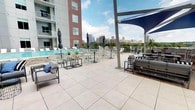 This modern urban residence offers a well-designed single-floor layout that balances contemporary style with functional living. The living area features warm hardwood flooring, large windows that invite abundant natural light, and a ceiling fan overhead. A tray ceiling detail adds architectural interest, while the open layout creates a comfortable, airy atmosphere with views of the surrounding cityscape.

The kitchen is outfitted with white shaker-style cabinetry, stainless steel appliances including a refrigerator, range, microwave, and dishwasher, complemented by dark countertops and a classic white subway tile backsplash. Open shelving adds both practicality and visual appeal, while hardwood flooring ties the space cohesively to the living area.

The bedroom is carpeted and generously sized, featuring dual windows with city views, a ceiling fan, and access to a closet and an adjoining bathroom. The bathroom features a combined tub and shower with a curved curtain rod, white cabinetry with a granite-style countertop, an undermount sink, and pendant lighting above a large mirror.

A standout feature is the expansive rooftop terrace, which offers panoramic city skyline views. The terrace is equipped with a pool, lounge chairs, dining and bar-height seating, shade sail structures, and comfortable outdoor sofas, providing an ideal setting for relaxation and social gatherings.