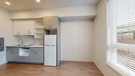 This efficiently designed 1-bedroom, 1-bathroom unit offers a thoughtful layout that maximizes every square foot. The open-concept living and kitchen area is anchored by large, double-pane windows fitted with horizontal blinds, flooding the space with natural light and offering views of the surrounding neighborhood. Light wood-tone laminate flooring runs throughout, complementing the clean, neutral color palette of warm whites and soft grays.

The kitchen is well-appointed with two-tone cabinetry — soft blue-gray lower cabinets paired with white upper cabinets — featuring sleek bar-pull hardware. A white quartz countertop provides ample prep space, while the kitchen includes a built-in microwave, cooktop, refrigerator, and a single-basin sink. Open shelving adds additional storage flexibility alongside the generous cabinet space.

The bedroom is a calm retreat, distinguished by a cool slate-blue accent wall that contrasts warmly with the neutral surroundings. Light switches and outlets are neatly positioned, and the room connects conveniently to the rest of the unit.

The bathroom is clean and functional, featuring a walk-in shower with a white surround and chrome fixtures, a toilet, and a wall-mounted vanity with a sink, all finished with the same light hardwood-style flooring found throughout the unit.