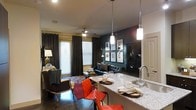 This single-floor space showcases an open-concept layout that connects the kitchen and living area into one cohesive, well-designed environment. The kitchen features a granite-topped island with seating, pendant lighting, and a modern faucet, making it both functional and visually striking. Vibrant red and burgundy bar chairs add a bold contrast against the neutral tones of the countertop. The living area is anchored by a dark leather sofa, layered with decorative pillows, and positioned beneath a gallery wall of framed artwork mounted against a deep teal accent wall. A dark display cabinet with glass paneling adds architectural interest and storage. Dark curtain panels frame the windows, which allow natural light to filter into the space, complementing the ceiling fan overhead. The flooring throughout appears to be a dark hardwood or polished concrete, contributing to the space's modern aesthetic. A patterned area rug defines the seating zone, while an upholstered armchair adds additional comfort. The overall ambiance is bold, curated, and contemporary, with a cohesive color palette of deep tones balanced by warm accent pieces.