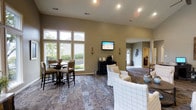 Located at 4800 Lake Forest Blvd in Westerville, Ohio, this single-floor community space offers a welcoming and well-appointed environment designed for comfort and social gathering. The open-concept layout seamlessly connects a lounge area with a casual dining space, creating a versatile setting for residents and guests alike.

The lounge area is furnished with plush, upholstered seating and a central coffee table, anchored by a wall-mounted flat-screen television flanked by decorative sconces. A beverage station nearby adds a convenient amenity for visitors. The dining area features high-top tables and chairs positioned beside a generous bank of windows that flood the space with natural light and offer views of the surrounding landscaping.

Soaring vaulted ceilings lend an airy, expansive feel throughout, while warm, neutral-toned walls and a mix of patterned carpet and hardwood-style flooring add texture and visual interest. Recessed lighting complements the natural light to keep the space bright and inviting. A connecting hallway leads to additional rooms, suggesting further amenity spaces beyond the main gathering area. Overall, the space strikes a balance between casual comfort and functional design, making it an ideal communal hub.