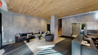 The lobby at 45 Main Street in Yonkers, NY is a thoughtfully designed contemporary entrance space that combines warm natural materials with sleek, modern finishes. The most striking architectural element is the expansive natural wood plank ceiling, which spans the entire lobby and introduces an organic warmth that contrasts beautifully with the large-format concrete wall panels lining the space.

Upon entry through the glass revolving door, residents and guests are greeted by an abundance of natural light streaming through floor-to-ceiling windows that face the street. A polished concrete concierge desk serves as the functional centerpiece, backed by a wood-clad accent wall featuring a built-in display shelf. A luggage cart stationed nearby underscores the building's full-service amenities.

The lounge area offers generous sectional seating in neutral tones, accented with teal and navy throw pillows, arranged around low circular side tables — providing a comfortable and inviting space for residents to gather. Recessed and track lighting complement the natural light throughout.

Dual stainless steel elevators are set against a concrete-paneled wall alongside an open staircase with natural wood treads, maintaining the lobby's cohesive material palette. The overall ambiance is refined, modern, and welcoming, reflecting a high standard of residential design in downtown Yonkers.
