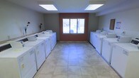 This single-floor shared laundry facility is designed for functionality and ease of use. The room is laid out symmetrically, with rows of full-sized top-loading washers and front-loading dryers lining both the left and right walls, providing ample capacity for multiple users simultaneously. The floor is covered in large, neutral-toned ceramic tiles that are easy to maintain and contribute to the clean, practical aesthetic of the space. Overhead, two large flush-mounted fluorescent light panels provide bright, even illumination throughout the room. A centrally positioned sliding window, framed by a terracotta-colored accent wall, serves as the room's focal point and allows natural light to supplement the overhead lighting while offering a view of the exterior. The combination of the light tile flooring and dual lighting sources creates a well-lit, open atmosphere. The layout provides a clear central walkway between the two banks of machines, allowing for comfortable movement and access. Overall, the space is efficiently organized and well-equipped to serve as a communal laundry area.