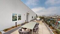Located at 1855 Westwood Blvd in Los Angeles, this rooftop terrace offers an expansive outdoor retreat with sweeping panoramic views of the Los Angeles skyline, including recognizable landmarks and the surrounding Westside neighborhoods. The terrace is finished with large-format tile flooring throughout, lending a clean and contemporary aesthetic to the open-air space.

A fully equipped outdoor kitchen anchors one end of the terrace, featuring a stainless steel built-in BBQ grill set into a sleek countertop surface — ideal for entertaining. Adjacent to the grill station, a dining area provides a rectangular table with comfortable woven rope chairs, accommodating a generous group for al fresco meals.

On the opposite end, a dedicated lounge zone features cushioned woven chairs positioned to take full advantage of the unobstructed city views. Glass panel railings line the perimeter of the terrace, ensuring safety while preserving the visual connection to the cityscape below.

Integrated raised planters filled with ornamental shrubs and young trees are distributed throughout the terrace, introducing greenery and softening the architectural lines of the surrounding white stucco building walls. The space is well-lit with low-profile bollard lighting and benefits from the open Los Angeles sky, making it equally suited for daytime gatherings or evening events.