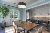 Located at 1400 Mall of Georgia Blvd in Buford, Georgia, this thoughtfully designed single-floor space offers a welcoming and functional environment ideal for meetings, gatherings, or collaborative use. The interior is finished with rich, dark hardwood flooring that contrasts elegantly with soft blue-gray walls and crisp white crown molding and trim throughout.

Natural light pours in through multiple large windows dressed with sheer white curtains, offering views of lush greenery outside. A central conference-style area features a substantial wood dining table surrounded by mid-century modern chairs, anchored by a woven jute area rug and illuminated by a striking black drum pendant light. A wall-mounted flat-screen display adds functionality to this primary gathering zone.

Adjacent to the main seating area, a secondary lounge space provides smaller round tables paired with dark accent chairs, creating an inviting setting for informal conversation. A glass-paned French door connects the interior to an exterior area, further enhancing the open, airy feel of the space.

A built-in banquette seating area with upholstered cushions and paired wall-mounted sconce lighting offers an additional intimate nook. Abstract artwork adorns one of the accent walls, adding a contemporary artistic touch. Together, these elements create a cohesive, refined ambiance that balances comfort with modern style.