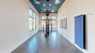 This single-floor Fitness On Demand studio is designed to provide a well-equipped, visually dynamic workout environment. The space is defined by its striking teal-painted ceiling, which contrasts sharply with the crisp white walls and creates a bold, energizing atmosphere. Recessed lighting and ceiling fans are integrated into the teal ceiling, ensuring the space remains well-lit and comfortable during workouts.

The flooring throughout features a herringbone-patterned tile in a cool gray tone, adding a sleek, modern aesthetic underfoot. Large windows along the perimeter flood the space with abundant natural light while offering views of the surrounding landscaped grounds and neighboring buildings.

A central multi-functional cable and resistance training rig serves as the focal point of the main workout area, accommodating a wide range of strength and functional fitness exercises. Stability balls are neatly stored near the entrance, alongside a wall-mounted yoga mat rack and a small touchscreen panel for on-demand class access. A large flat-screen television mounted on the teal accent wall displays the Fitness On Demand platform, enabling users to follow guided workout programs. The layout is open and uncluttered, allowing ample space for movement and exercise.