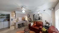 This single-floor residence offers an inviting open-concept layout that seamlessly connects the living area to the kitchen, creating a cohesive and functional living environment. The living room is warmly appointed with plush carpeting, a bold leather sofa accented with colorful throw pillows, and eye-catching wall décor including a striking zebra-print canvas and a cluster of decorative mirrors. A ceiling fan with a built-in light fixture anchors the room, providing both comfort and illumination. The kitchen is efficiently designed with light-toned cabinetry, a smooth countertop, and a peninsula breakfast bar with seating for two, making it ideal for casual dining or entertaining. Appliances are integrated neatly into the layout, contributing to a clean and organized aesthetic. A textured popcorn ceiling runs throughout, adding a classic residential character. The space benefits from a thoughtful blend of ambient and accent lighting, including a sleek multi-arm floor lamp in the living area. Overall, the residence presents a cozy yet stylish atmosphere, balancing practical design with personalized décor touches throughout its single level.