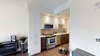 This single-floor residence showcases a thoughtfully designed contemporary interior with warm wood-tone flooring that flows consistently throughout the space. The kitchen is efficiently organized within a defined alcove, featuring a two-tone cabinet scheme that pairs crisp white upper cabinetry with darker lower cabinets. Stainless steel appliances—including a range, over-the-range microwave, and dishwasher—add a polished finish, while a neutral tile backsplash and recessed lighting complete the functional yet refined aesthetic. A tall floor-to-ceiling cabinet column provides generous storage just outside the kitchen entry.

The bedroom is a striking space anchored by a bold black accent wall adorned with a gallery arrangement of black-and-white artwork. A seating area with a sectional sofa and gold accent table adds versatility, while a large window with sheer draping offers views of the surrounding cityscape and fills the room with natural light. The bedroom's neutral palette is complemented by layered textiles and contemporary furnishings.

The bathroom maintains the residence's clean aesthetic with full-height tile surrounding a soaking tub and shower combination, chrome fixtures, and wood-look flooring. Overall, the space presents a cohesive modern design with a neutral, urban-inspired palette.