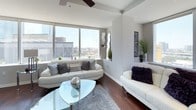 This two-bedroom, two-bathroom residence offers a sophisticated urban living experience with a cohesive modern design aesthetic throughout. Dark hardwood flooring flows continuously across the entire unit, anchoring each space with warmth and contrast against the light, neutral-toned walls.

The living area is a standout space, featuring expansive floor-to-ceiling windows that frame impressive city skyline views and flood the room with natural light. A ceiling fan adds functionality, while the open layout connects seamlessly to the dining area and kitchen.

The kitchen is outfitted with dark espresso cabinetry, stainless steel appliances including a full-size refrigerator, microwave, and range, as well as a marble-style countertop island. Track lighting above enhances the contemporary feel, and an adjacent glass-top dining table with modern seating completes the space.

The primary bedroom features city views through large windows, a ceiling fan, and direct access to an en-suite bathroom equipped with dark cabinetry, marble countertops, and a tiled shower. The second bedroom is comfortably sized with its own ceiling fan. The second bathroom mirrors the first in finish quality, offering a tub-shower combination with white tile surround.