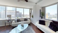 This two-bedroom, two-bathroom residence offers a sophisticated urban living experience with a cohesive modern design aesthetic throughout. Dark hardwood flooring flows continuously across the entire unit, anchoring each space with warmth and contrast against the light, neutral-toned walls.

The living area is a standout space, featuring expansive floor-to-ceiling windows that frame impressive city skyline views and flood the room with natural light. A ceiling fan adds functionality, while the open layout connects seamlessly to the dining area and kitchen.

The kitchen is outfitted with dark espresso cabinetry, stainless steel appliances including a full-size refrigerator, microwave, and range, as well as a marble-style countertop island. Track lighting above enhances the contemporary feel, and an adjacent glass-top dining table with modern seating completes the space.

The primary bedroom features city views through large windows, a ceiling fan, and direct access to an en-suite bathroom equipped with dark cabinetry, marble countertops, and a tiled shower. The second bedroom is comfortably sized with its own ceiling fan. The second bathroom mirrors the first in finish quality, offering a tub-shower combination with white tile surround.
