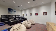This dedicated screening room is thoughtfully designed to deliver an immersive entertainment experience. The space is arranged with two tiers of seating: a raised row of plush black leather recliner chairs with built-in armrests and cup holders occupies the elevated rear section, while oversized bean bag chairs offer a more relaxed viewing option at the front. Blue LED strip lighting accents the tiered step, adding a cinematic atmosphere and serving as practical aisle lighting.

The room is finished with crisp white walls and recessed ceiling lighting, complemented by flush-mounted in-ceiling speakers that suggest a robust audio setup. A large flat-screen television is mounted on the primary viewing wall, accompanied by a slatted wood media console beneath it for equipment storage.

The décor is a distinctive feature of the room, with a series of bold, colorful pop art portraits displayed along the side walls, alongside an abstract blue canvas painting positioned prominently above the seating area. The flooring throughout is a patterned carpet in dark tones with red accents, reinforcing the theatrical aesthetic. A signage panel near the entrance provides room guidelines, indicating this is a shared or amenity-style space designed for group use.