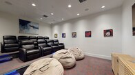 This dedicated screening room is thoughtfully designed to deliver an immersive entertainment experience. The space is arranged with two tiers of seating: a raised row of plush black leather recliner chairs with built-in armrests and cup holders occupies the elevated rear section, while oversized bean bag chairs offer a more relaxed viewing option at the front. Blue LED strip lighting accents the tiered step, adding a cinematic atmosphere and serving as practical aisle lighting.

The room is finished with crisp white walls and recessed ceiling lighting, complemented by flush-mounted in-ceiling speakers that suggest a robust audio setup. A large flat-screen television is mounted on the primary viewing wall, accompanied by a slatted wood media console beneath it for equipment storage.

The décor is a distinctive feature of the room, with a series of bold, colorful pop art portraits displayed along the side walls, alongside an abstract blue canvas painting positioned prominently above the seating area. The flooring throughout is a patterned carpet in dark tones with red accents, reinforcing the theatrical aesthetic. A signage panel near the entrance provides room guidelines, indicating this is a shared or amenity-style space designed for group use.