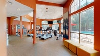 Located at 3301 Glenn McConnell Pkwy in Charleston, South Carolina, this single-floor fitness center is a well-equipped and visually striking facility designed to energize and motivate. The space is defined by bold, vibrant orange walls that lend a dynamic and high-energy atmosphere throughout. The layout is divided into distinct functional zones, seamlessly connected to create a comprehensive workout environment.

The cardio area features a vaulted, octagonal ceiling with recessed lighting and a ceiling fan, creating an airy, open feel. Floor-to-ceiling arched windows and large glass panels flood the space with abundant natural light and provide views of the outdoor pool and surrounding greenery. Multiple treadmills and elliptical machines are arranged to face the windows, enhancing the workout experience. Wall-mounted televisions offer additional entertainment.

A dedicated strength and free weights section includes cable machines, adjustable benches, weight racks with dumbbells, and various resistance equipment. Full-length mirrors line the walls, amplifying the sense of space and supporting proper form during exercise. Ceiling fans maintain comfortable airflow throughout.

A transitional lounge area with modular seating connects the fitness zones and opens through glass sliding doors to adjacent interior spaces. The combination of natural light, thoughtful equipment layout, and energizing design makes this fitness center a standout amenity.