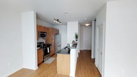 This single-floor residence showcases a thoughtfully designed open-concept kitchen that serves as the heart of the space. The kitchen is outfitted with a full suite of stainless steel appliances, including a French door refrigerator, electric range, and built-in microwave, all complemented by rich warm-toned wood cabinetry that extends to the ceiling for maximized storage. Dark granite countertops provide a striking contrast against the lighter elements of the space, continuing onto a peninsula that doubles as a breakfast bar and defines the boundary between the kitchen and the adjoining hallway.

The flooring throughout is light-toned hardwood, lending warmth and continuity to the layout. The entryway is visible from the kitchen, featuring a closet with louvered doors for convenient storage. Recessed and track lighting illuminate the kitchen area effectively, while flush-mount fixtures carry the lighting through the hallway. The overall color palette is neutral, with crisp white walls and ceilings creating a clean, bright atmosphere. The layout is functional and efficient, with a clear sightline from the kitchen through to the entry.