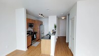 This single-floor residence showcases a thoughtfully designed open-concept kitchen that serves as the heart of the space. The kitchen is outfitted with a full suite of stainless steel appliances, including a French door refrigerator, electric range, and built-in microwave, all complemented by rich warm-toned wood cabinetry that extends to the ceiling for maximized storage. Dark granite countertops provide a striking contrast against the lighter elements of the space, continuing onto a peninsula that doubles as a breakfast bar and defines the boundary between the kitchen and the adjoining hallway.

The flooring throughout is light-toned hardwood, lending warmth and continuity to the layout. The entryway is visible from the kitchen, featuring a closet with louvered doors for convenient storage. Recessed and track lighting illuminate the kitchen area effectively, while flush-mount fixtures carry the lighting through the hallway. The overall color palette is neutral, with crisp white walls and ceilings creating a clean, bright atmosphere. The layout is functional and efficient, with a clear sightline from the kitchen through to the entry.
