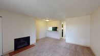 This single-story residence offers a practical and functional layout spread across a series of well-defined living spaces. The living room serves as the central gathering area, featuring neutral-toned wall-to-wall carpeting, cream-colored walls, and a textured ceiling. A wood-burning fireplace with a tile hearth anchors one wall, adding a cozy focal point to the room. A pass-through window connects the living area to the kitchen, promoting an open feel and convenient interaction between spaces.

The kitchen is equipped with a black electric range, an overhead ventilation hood, warm wood-toned cabinetry, and granite-style countertops, providing both functionality and a degree of visual warmth. The bedroom features soft carpeting, a textured ceiling, baseboard heating, a wall-mounted air conditioning unit, and a window that allows natural light and outdoor views into the space.

The bathroom is straightforward in design, including a full-size tub and shower combination with white tile surround, a toilet, and wood-style flooring. Throughout the unit, baseboard heating provides consistent climate control. The overall aesthetic is clean, neutral, and move-in ready, offering a comfortable and low-maintenance living environment.