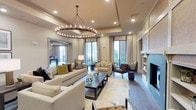 This single-floor space showcases a thoughtfully designed lounge or common area that exudes refined elegance. The room's focal point is a striking circular chandelier with candle-style lights suspended from a tray ceiling, casting warm, inviting light throughout. Neutral tones dominate the palette, with cream and beige walls and furnishings accented by bold pops of navy, chartreuse, and gold through decorative pillows and accessories.

The generous seating arrangement includes plush sofas and accent chairs arranged around a dark wood coffee table atop a light-toned area rug, creating distinct yet open conversation zones. A built-in fireplace with a clean, contemporary surround anchors one wall, flanked by integrated shelving. Tall windows dressed with floor-length drapes allow substantial natural light and offer views of an exterior courtyard, enhancing the sense of openness. Dark hardwood flooring grounds the space with warmth and contrast.

A large framed mirror near the entryway amplifies depth and light, while recessed lighting supplements the chandelier for layered illumination. The overall ambiance is polished, welcoming, and carefully curated.