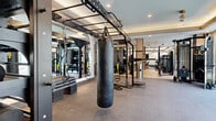 Located at 518 W 8th St in Dallas, TX, this contemporary fitness center offers a generously sized, single-floor layout designed to accommodate a full range of workout disciplines. The space is divided into two distinct zones that transition seamlessly from one to the next.

The first zone features light hardwood-style flooring, tall ceilings with sleek linear pendant lighting, and large windows that invite abundant natural light and views of the surrounding neighborhood. This area is outfitted with Matrix cable machines, multi-functional strength stations, and adjustable benches, providing a clean and modern environment for resistance training.

The second zone transitions to durable rubber flooring and is anchored by a tray ceiling with recessed lighting and ceiling fans, creating a comfortable atmosphere for high-intensity training. Floor-to-ceiling windows line one wall, flooding the space with daylight. This area houses a TRX suspension training rig, a heavy punching bag, additional cable and strength machines, and a full row of cardio equipment including treadmills, ellipticals, and stationary bikes. Full-length mirrors amplify the sense of openness throughout. A water fountain and additional amenities are accessible from within the space, rounding out this well-appointed fitness facility.