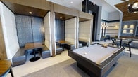 This contemporary amenity lounge is designed to offer both social engagement and relaxed retreat within a single, thoughtfully appointed floor. The space centers around a sleek pool table finished in light felt with dark wood trim, serving as a natural focal point for recreation and casual gatherings. Flanking the gaming area is a row of private booth-style seating nooks, each upholstered in plush cream and gray tufted panels and separated by warm wood partitions. These semi-enclosed booths feature individual tables, creating intimate settings ideal for small group conversations or quiet work sessions. The textured wall treatments within each nook add depth and visual interest, complementing the overall refined aesthetic. Overhead, recessed lighting provides a warm, ambient glow that enhances the sophisticated atmosphere. The flooring transitions between soft carpet and smooth hard surface, subtly delineating activity zones within the open layout. In the background, a mirrored wall expands the sense of space, reflecting pendant lighting fixtures that add an elegant touch. The overall design balances modern luxury with functional versatility, making this lounge a compelling destination for both leisure and socializing.