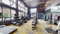 This contemporary fitness center is designed to deliver a premium workout experience through both its thoughtful layout and high-end equipment selection. The space features soaring ceilings fitted with sleek linear pendant lighting and ceiling fans, creating an open, airy atmosphere that feels energizing rather than confined. Floor-to-ceiling windows line the perimeter walls, flooding the interior with natural light and offering views of an outdoor amenity area, including a patio with seating and landscaped greenery.

The flooring makes an immediate visual impression — bold yellow and grey geometric-patterned carpet tiles lend the space a modern, dynamic character that complements the dark charcoal wall palette throughout.

Equipment is thoughtfully organized across the floor. A dedicated cardio zone houses a lineup of True-brand treadmills, elliptical cross-trainers, upright and recumbent bikes, and a stair climber. The strength training area features a multi-station cable and pulley system, a functional trainer, adjustable benches, and a rack with free weights and medicine balls. Circular mirror fixtures mounted on the walls add a contemporary design element while enhancing spatial depth. The overall ambiance is sleek, motivating, and well-suited for a full-body workout routine.