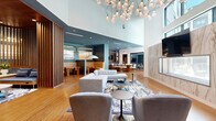Located at 5851 Columbia Pike in Falls Church, Virginia, this sophisticated amenity space spans a generous single floor and is designed to impress with a seamless blend of elegance and functionality. The centerpiece is a soaring double-height lounge area bathed in natural light from expansive windows, anchored by a striking sculptural pendant chandelier and warm hardwood flooring. A dramatic marble accent wall paired with a mounted display and custom wall art adds a refined artistic touch to the main gathering area. Plush sectional sofas, accent chairs, and patterned area rugs create inviting conversation zones throughout the open layout. A richly wood-paneled nook with upholstered bench seating and a marble-topped coffee table offers an intimate retreat within the larger space. The kitchen and bar area features a bold teal palette, bar-height seating, and built-in cabinetry, ideal for entertaining or casual gatherings. Three private offices provide focused work environments, one of which boasts floor-to-ceiling glass walls overlooking an outdoor courtyard with lush greenery. An additional meeting room with a round conference table and integrated display screen supports collaborative work. The entrance and corridor areas maintain the design continuity with clean lines, curated greenery, and abundant natural light, delivering a polished, resort-inspired atmosphere throughout.