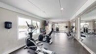 Located at 2500 E James Ave in Baytown, Texas, this fitness center offers a thoughtfully designed exercise space that accommodates a full range of workout needs. The elongated, single-floor layout is bright and open, with large windows lining multiple walls that flood the room with natural light and provide views of the surrounding outdoor areas, including a pool and lush landscaping.

The durable, rubber-textured flooring provides both safety and comfort underfoot, ideal for high-activity use. The space is climate-controlled with a wall-mounted split unit, ensuring a comfortable environment year-round. A large flat-screen television is mounted on a whitewashed brick accent wall, adding a functional entertainment element to the workout experience.

Cardio equipment includes Spirit-brand treadmills, elliptical trainers, and a recumbent bike, all positioned to take advantage of the natural light and outdoor views. The strength training section features a multi-function cable machine, a free weight rack with a full set of dumbbells, a weight bench, and a medicine ball rack with multiple weighted balls. Floor-to-ceiling mirrors along one wall enhance the sense of space and allow users to monitor their form. The fitness center is accessed through double glass-paned doors and is designed to support a complete and effective workout routine.