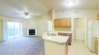 This single-floor residence is designed with an open-concept layout that seamlessly connects the kitchen and living areas. The kitchen features a wraparound peninsula with a built-in double sink and light countertops, providing functional workspace and a natural division between the cooking and living zones. Warm honey-toned wood cabinetry lines the upper walls, offering ample storage, while white appliances — including a refrigerator, microwave, and range — are neatly integrated beneath. A pendant light fixture adds a subtle decorative touch above the peninsula.

The adjacent living area is carpeted in a neutral tone and anchored by a white-mantled fireplace, creating a welcoming focal point. Vaulted ceilings with a ceiling fan enhance the sense of openness and airflow throughout the space. Soft, neutral wall tones contribute to a calm and cohesive atmosphere. A sliding glass door at the far end of the living area allows natural light to filter in and provides access to the exterior. Tile flooring transitions from the kitchen area, while plush carpet covers the living space, balancing comfort with practicality throughout this well-configured home.