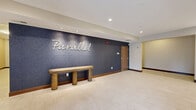 The lobby of Parallel, located at 1415 County Road B W in Roseville, Minnesota, serves as an elegant and functional gateway to the building. The single-floor space is finished with large-format light tile flooring throughout, creating a clean and cohesive aesthetic that flows seamlessly from the entrance to the interior hallway areas.

Upon entry through glass automatic doors, residents are greeted by a prominent navy blue textured feature wall bearing the scripted "Parallel" logo, accompanied by a solid wood bench beneath it — a simple yet stylish focal point. Recessed lighting illuminates the space evenly, enhancing the bright, open feel of the interior.

Adjacent to the entrance, a built-in credenza houses a coffee station and interactive kiosk, offering convenient amenities for daily use. A round mirror adds a decorative touch to this area.

Deeper into the lobby, a dedicated mail and package room features rows of secure mailboxes flanking a central wooden island workstation. This area is distinguished by a backlit textured tile accent wall and a wood-slat ceiling detail overhead, adding warmth and architectural interest. Vertical linear light fixtures further enhance the refined design of this functional space.

Rich dark wood base trim ties all areas together, complementing the warm wood tones of the furnishings and creating a polished, contemporary atmosphere throughout.