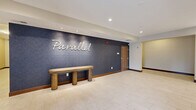 The lobby of Parallel, located at 1415 County Road B W in Roseville, Minnesota, serves as an elegant and functional gateway to the building. The single-floor space is finished with large-format light tile flooring throughout, creating a clean and cohesive aesthetic that flows seamlessly from the entrance to the interior hallway areas.

Upon entry through glass automatic doors, residents are greeted by a prominent navy blue textured feature wall bearing the scripted "Parallel" logo, accompanied by a solid wood bench beneath it — a simple yet stylish focal point. Recessed lighting illuminates the space evenly, enhancing the bright, open feel of the interior.

Adjacent to the entrance, a built-in credenza houses a coffee station and interactive kiosk, offering convenient amenities for daily use. A round mirror adds a decorative touch to this area.

Deeper into the lobby, a dedicated mail and package room features rows of secure mailboxes flanking a central wooden island workstation. This area is distinguished by a backlit textured tile accent wall and a wood-slat ceiling detail overhead, adding warmth and architectural interest. Vertical linear light fixtures further enhance the refined design of this functional space.

Rich dark wood base trim ties all areas together, complementing the warm wood tones of the furnishings and creating a polished, contemporary atmosphere throughout.
