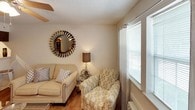 This single-floor residence offers a well-organized layout across several functional spaces, all unified by warm hardwood-style flooring and textured ceilings throughout. The living area provides a comfortable and intimate setting, featuring a window with blinds and sheer curtains that allow natural light to filter in, complemented by a ceiling fan that enhances airflow. A decorative round mirror accents the neutral-toned walls, adding visual interest to the space.

The bedroom is adequately sized to accommodate standard furnishings and includes a ceiling fan, a window for natural light, and a closet door for storage access. A walk-in closet, visible from the main living area, offers well-organized shelving and generous storage capacity.

The kitchen is efficiently designed with dark wood cabinetry, light countertops, a white refrigerator, an electric range, and a sink — providing all essential cooking amenities in a compact galley-style layout. A breakfast bar with metal stools connects the kitchen to the adjacent living space, creating a casual dining option.

The bathroom features a single-vanity with a wide mirror, a lighted fixture above, and a tub-shower combination with a curtain, all finished in clean, neutral tones.