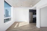 This single-floor unit showcases a clean, modern aesthetic throughout, with light hardwood-style flooring and crisp white walls creating a bright, cohesive atmosphere. The kitchen is well-appointed with flat-front grey cabinetry, white quartz countertops, a subway tile backsplash, stainless steel appliances including a gas range, built-in microwave, and dishwasher, complemented by a deep stainless steel sink with a contemporary pull-down faucet. A narrow hallway leads to a dedicated laundry area featuring a stacked washer and dryer unit, adding everyday convenience. The bathroom offers a full tub-shower combination with large-format white tile surround, chrome fixtures, and neutral tile flooring. The living and sleeping areas are bathed in abundant natural light through expansive floor-to-ceiling windows that frame sweeping urban skyline views, reflecting the unit's elevated position within the building. Exposed concrete ceilings in the main rooms add an industrial design contrast to the otherwise light and airy interior, lending the space a distinctly contemporary character throughout.