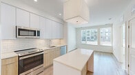 Located at 8005 Gramercy Blvd in Rockville, MD, this single-floor residence offers a thoughtfully designed layout with modern finishes and a bright, airy ambiance. The kitchen is a standout feature, showcasing a clean contemporary aesthetic with two-tone cabinetry — crisp white upper cabinets paired with warm natural wood lower cabinets — set against a classic white subway tile backsplash. Stainless steel appliances, including a refrigerator, electric range, built-in microwave, and dishwasher, complement the sleek quartz countertops. A central island provides additional prep and dining space, while a large pendant light fixture anchors the room. Light hardwood flooring flows seamlessly throughout the unit, enhancing the open and cohesive feel. Living areas benefit from generous natural light through oversized windows, offering views of the surrounding community. The bathroom is finished with large-format stone-look tile on both the floors and shower surround, paired with a wood-toned vanity and a full soaking tub. Bedrooms are spacious and bright, with neutral walls and natural light streaming in through well-positioned windows. The overall design palette — white, natural wood, and soft neutral tones — creates a clean, modern, and welcoming atmosphere.