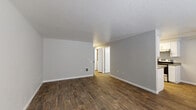 Located at 700 SW Eastman Pkwy in Gresham, Oregon, this single-floor residence offers a well-organized layout with a clean, modern aesthetic throughout. The open living area features rich, dark wood-look flooring and neutral gray walls, creating a warm yet contemporary atmosphere. A wall-mounted heating unit and ample wall space make this room both functional and adaptable. The living area flows naturally toward the kitchen, which is efficiently designed with white cabinetry, gray countertops, stainless steel appliances including a refrigerator and electric range, and light wood-look flooring that ties the space together cohesively. A dedicated entry area with a front-facing window allows natural light to fill the space. The carpeted bedrooms provide a softer, quieter retreat, with one room featuring a sliding glass door fitted with vertical blinds that opens to an outdoor view, and another room with a window overlooking the surrounding greenery. The bathroom is finished with wood-look tile flooring, a wide vanity with dual sinks, gray countertops, white cabinetry, and a well-lit mirror — offering both style and practicality. Textured ceilings are consistent throughout, adding a classic touch to the overall interior character.