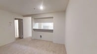 This single-floor residence offers a clean, understated interior designed for comfortable everyday living. The living area is finished with soft, neutral-toned carpeting that lends warmth underfoot, while the crisp white walls and recessed lighting create a bright, airy atmosphere. A standout architectural feature is the built-in pass-through window that connects the living space to the kitchen, framed neatly within the wall and topped with a countertop ledge — ideal for casual dining, serving, or simply maintaining an open sightline between the two areas. The kitchen, visible through the pass-through, is outfitted with white cabinetry and appliances, maintaining the cohesive, clean palette found throughout the home. The layout is straightforward and efficient, making good use of the available space without unnecessary complexity. A doorway visible to one side suggests additional rooms or storage areas accessible from the main living space. The overall ambiance is neutral and move-in ready, providing a versatile blank canvas well-suited for a variety of interior styles and furnishing choices. The consistent use of light tones across flooring, walls, and cabinetry gives the entire space a unified, polished feel.