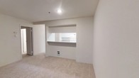 This single-floor residence offers a clean, understated interior designed for comfortable everyday living. The living area is finished with soft, neutral-toned carpeting that lends warmth underfoot, while the crisp white walls and recessed lighting create a bright, airy atmosphere. A standout architectural feature is the built-in pass-through window that connects the living space to the kitchen, framed neatly within the wall and topped with a countertop ledge — ideal for casual dining, serving, or simply maintaining an open sightline between the two areas. The kitchen, visible through the pass-through, is outfitted with white cabinetry and appliances, maintaining the cohesive, clean palette found throughout the home. The layout is straightforward and efficient, making good use of the available space without unnecessary complexity. A doorway visible to one side suggests additional rooms or storage areas accessible from the main living space. The overall ambiance is neutral and move-in ready, providing a versatile blank canvas well-suited for a variety of interior styles and furnishing choices. The consistent use of light tones across flooring, walls, and cabinetry gives the entire space a unified, polished feel.
