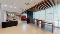 This contemporary single-floor space serves as a stylish communal lounge and lobby area, thoughtfully designed to balance openness with defined zones for gathering and relaxation. The layout flows seamlessly across light-toned tile and hardwood flooring, creating a welcoming and cohesive environment throughout.

A striking architectural feature is the slatted wood ceiling installation, which adds warmth and visual depth while delineating the dining and lounge zones. Floor-to-ceiling sheer curtains line the expansive glazed walls, diffusing abundant natural light and providing soft views of the greenery outside. Pendant lighting and adjustable track spotlights complement the natural illumination, ensuring a well-lit atmosphere at all times.

The seating areas are diverse and thoughtfully arranged. A reception or concierge desk anchors one section of the space, accompanied by intimate lounge clusters featuring low barrel chairs in dark tones with bold yellow accents. A raised dining area offers long communal high-top tables with vibrant orange bar seating, while round café-style tables provide additional casual options. A built-in banquette with red upholstery and patterned cushions adds a pop of color to one corner.

Curated contemporary artwork adorns the walls, reinforcing the space's modern aesthetic. Dark navy accent walls provide dramatic contrast against the otherwise light and airy palette. The overall ambiance is polished, energetic, and inviting.
