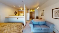 Located at 1702 Dublin Trail in Neenah, Wisconsin, this single-floor unit offers a well-organized, open-concept layout that seamlessly connects its key living spaces. The kitchen is outfitted with light maple cabinetry, warm yellow countertops, a full suite of appliances including an electric range with overhead ventilation, a refrigerator, and ample storage both above and below the counters. A half-wall pass-through connects the kitchen to the main living area, enhancing the sense of openness throughout. The living area features neutral carpet flooring, recessed and track lighting, and space for seating and a dining area. Framed artwork adorns the walls, adding a finished, welcoming character to the room. The bedroom is carpeted and benefits from a large window fitted with vertical blinds that allow for natural light while maintaining privacy. A full bathroom includes a shower and tub combination with a curtain, a wood-toned vanity with an integrated sink, a large mirror, and tile flooring. Throughout the unit, the color palette remains light and neutral, contributing to a clean, practical atmosphere. The layout, as visible in the dollhouse view, is compact yet efficiently arranged, making thoughtful use of the available floor space.