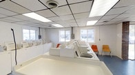 Located at 4301 S 6th St in Terre Haute, Indiana, this laundromat occupies a single-floor commercial space designed for efficiency and ease of use. The interior features a clean, open layout with rows of top-load washing machines arranged in a structured configuration, providing ample capacity for customers. The facility is well-lit with recessed fluorescent panel lighting set within a drop-tile ceiling, ensuring a bright and comfortable environment throughout the day. Multiple windows along the exterior walls allow natural light to supplement the overhead lighting, offering views of the surrounding area and contributing to an open, airy atmosphere. The walls are finished in a crisp white, reinforcing the clean and sanitary nature of the space. Seating is provided along the machine rows in the form of sturdy chairs, giving patrons a convenient place to wait. Electrical panels and utility connections are visibly integrated into the walls, reflecting the functional, utilitarian design of the facility. The flooring is durable and easy to maintain, suited for the demands of a high-traffic commercial laundry environment. Overall, this laundromat presents a practical, no-frills space equipped to serve the laundry needs of the surrounding Terre Haute community.