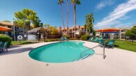 Located at 8401 E 22nd St in Tucson, Arizona, this outdoor amenity space serves as the communal hub of a well-established residential community. The centerpiece is a generously sized, kidney-shaped swimming pool with crystal-clear turquoise water, surrounded by a smooth, light-colored concrete pool deck. Turquoise lounge chairs are arranged along the poolside, inviting residents to relax and enjoy Tucson's abundant sunshine. Shaded dining areas with colorful market umbrellas provide comfortable spots for outdoor gatherings. Towering palm trees and mature shade trees frame the space, lending a resort-like atmosphere that is characteristic of the Southwest. Adjacent to the pool area is a well-maintained courtyard featuring desert-friendly landscaping with decorative gravel, low-maintenance plantings, and a turf seating area outfitted with Adirondack chairs — ideal for casual outdoor socializing. The surrounding multi-story buildings are rendered in a classic Southwestern stucco style with warm earth tones and tiled roof accents, reinforcing the regional architectural character. Iron fencing encloses the amenity areas, providing both security and visual definition. The overall ambiance is relaxed, open, and perfectly suited to the sunny Tucson climate.