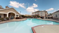 Located at Amelia Station in Clayton, North Carolina, this single-level outdoor amenity space is designed to deliver a resort-style experience for residents. At its centerpiece is a generously sized, freeform swimming pool with a classic brick coping edge, surrounded by an expansive concrete pool deck lined with lounge chairs—ideal for relaxing under the Carolina sun.

A covered pavilion anchors the space, providing shaded outdoor living with several distinct seating areas. A striking floor-to-ceiling stone fireplace with a rustic wood mantle and branded "A" signage serves as a focal point, flanked by comfortable wicker seating with teal cushions. Adjacent to the fireplace, an outdoor bar counter finished with a dark granite top and white paneling offers bar-height wicker seating and is equipped with two wall-mounted flat-screen TVs, creating an ideal spot for social gatherings.

Additional dining areas beneath the covered pavilion feature round metal tables with wicker chairs, offering views of the pool. A separate open-air patio with umbrella shading and a built-in grill station provides further entertaining options. The entire area is enclosed by black iron fencing, with landscaped grounds and views of the surrounding residential community completing the inviting outdoor atmosphere.