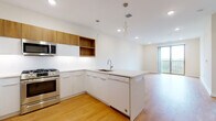 This single-floor residence showcases a clean, contemporary design highlighted by warm hardwood-style flooring that flows throughout the main living areas. The open-concept kitchen is a standout feature, equipped with stainless steel appliances including a gas range, built-in microwave, side-by-side refrigerator with ice maker, and dishwasher. Cabinetry combines white lacquered lower units with warm wood-toned upper cabinets and open shelving, complemented by white countertops and a subway tile backsplash. A peninsula with pendant lighting separates the kitchen from the spacious living area, which benefits from natural light and direct access to a private balcony with open green views. The bedroom also features balcony access through a sliding glass door, with soft carpeting adding comfort underfoot. The bathroom is finished with large-format tile flooring, a wood-toned floating vanity with white countertop, and a glass-enclosed walk-in shower with white tile surround. A dedicated in-unit laundry area accommodates a stacked full-size washer and dryer, adding everyday convenience to this well-appointed space.