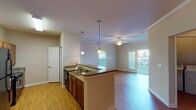 Located at 2000 Alexan Dr in Cary, North Carolina, this single-floor residence offers a well-designed open layout that seamlessly connects the kitchen, dining, and living areas. The kitchen is a functional centerpiece, featuring dark granite countertops, rich wood cabinetry, black appliances including a refrigerator, gas range, and dishwasher, and a peninsula with pendant lighting overhead. A double sink and ample counter space make it well-suited for everyday cooking and entertaining. Crown molding and ceiling fans accent the living and dining areas, which are bathed in natural light from large windows and a sliding glass door that opens to a private balcony with outdoor views. Hardwood-style flooring runs consistently throughout the main living spaces and bedroom, lending warmth and continuity to the interior. The bedroom is generously proportioned with vaulted ceilings, a ceiling fan, and a sidelight window by the entry door. The bathroom includes a soaking tub with tiled surround, a granite-topped vanity, and hardwood-style flooring. A dedicated laundry area houses a full-size washer and dryer. Track lighting in the dining space adds a modern touch to the otherwise classic, comfortable aesthetic of this Cary home.