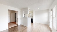This single-floor residence offers a clean, functional layout with a bright, neutral palette throughout. The living area is open and airy, featuring light wood-style flooring and a textured ceiling. Natural light flows in through slatted blinds on multiple windows, and a glass door leads to an exterior access point, enhancing the sense of openness. A half-wall with a pass-through window connects the living space to the kitchen, creating an interactive feel while maintaining visual separation.

The galley-style kitchen is efficiently arranged with white cabinetry lining both sides, light stone-look countertops, and black appliances including a range and refrigerator. A wall of upper cabinets provides ample storage, and wood-style flooring continues underfoot.

The bedroom is carpeted and features a window that admits natural light, contributing to a calm and comfortable atmosphere. White walls and clean finishes are consistent throughout.

The bathroom is well-appointed with a vanity featuring a stone-look countertop, single sink, and mirrored storage above. A notable feature is the stacked in-unit washer and dryer tucked neatly alongside the vanity, adding significant convenience. The space is cohesive, practical, and move-in ready.