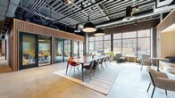 Located at 301 Winchester Avenue in New Haven, CT, this single-floor commercial space delivers a compelling blend of industrial aesthetics and modern workplace design. The layout spans a generous open-plan footprint, defined by polished concrete floors, exposed ductwork, and a dark ceiling grid that together create a distinctly urban, loft-inspired atmosphere. Large floor-to-ceiling windows line the perimeter, flooding the interior with natural light and offering views of the surrounding New Haven streetscape and neighboring brick buildings. A central conference area features a long communal table paired with a mix of molded and upholstered chairs, anchored by a light-toned area rug and illuminated by oversized pendant lights. Glass-walled private rooms with dark metal frames provide enclosed meeting and collaboration spaces without sacrificing visual openness. Warm vertical wood slat partitions add texture and acoustic softening throughout the space, contrasting beautifully with the industrial ceiling above. Additional seating zones include lounge chairs, intimate café-style tables, and built-in banquette nooks framed in natural wood. Lush potted greenery is strategically placed to add life and warmth. The overall ambiance is professional yet inviting, designed to support a variety of work styles.