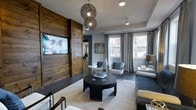 Located at 2170 Benton Blvd in Savannah, Georgia, this single-story space masterfully combines rustic charm with modern sophistication. The living area serves as the centerpiece of the space, anchored by a dramatic reclaimed wood accent wall that houses a large flat-screen display, creating a bold focal point that contrasts beautifully with the cool gray tones of the surrounding walls. Dark tile flooring grounds the space, transitioning seamlessly to a plush area rug that defines the seating arrangement. A round dark wood coffee table sits at the heart of the room, flanked by contemporary upholstered chairs with blue accent pillows and a deep gray sofa. Overhead, a pair of sculptural wire globe pendant lights add an artistic touch, complementing the recessed lighting throughout. Large windows dressed in flowing gray curtains invite generous natural light and offer views of the surrounding greenery, enhancing the open and airy feel of the interior. The overall ambiance strikes a refined balance between warmth and modernity, making this a welcoming and visually engaging space well-suited for both relaxation and everyday living.
