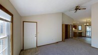 This single-floor space is characterized by its vaulted ceilings, which lend a sense of openness and volume to the main living area. The entryway, finished with tile flooring, transitions seamlessly into a carpeted living space, creating a clear delineation between arrival and relaxation zones. Neutral beige walls and white trim throughout contribute to a clean, understated aesthetic. A ceiling fan is mounted at the peak of the vaulted ceiling, providing both air circulation and a functional focal point overhead. The open layout allows a clear sightline from the front entry toward the rear of the unit, where a hallway leads to additional rooms. Wood-toned door frames and trim add warmth against the light-colored walls. Visible in the background, the kitchen features wood cabinetry with a warm finish and light-colored countertops, offering a functional cooking space. A corridor lined with closet doors provides practical storage solutions. The overall ambiance is straightforward and functional, with architectural details like the vaulted ceiling elevating the character of an otherwise modest, efficiently designed space.
