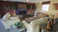 This single-floor lounge space exudes a warm and inviting ambiance, thoughtfully designed for comfort and community. The focal point of the room is a sleek, built-in fireplace framed by a bold, dark surround and flanked by decorative botanical artwork, set against deep burgundy accent walls that lend the space a rich, cozy character. The seating arrangement is generous and well-considered, featuring plush cream sofas accented with patterned throw pillows, complemented by woven rattan armchairs that add a touch of texture and warmth. Lush potted trees and a trailing ivy centerpiece bring natural greenery into the interior, softening the overall aesthetic. A tiled-top coffee table serves as a stylish and functional gathering point at the heart of the arrangement. Track lighting runs along the ceiling, providing adjustable illumination throughout the space, while large windows along one wall invite natural light and offer views to the exterior. The combination of layered textures, warm tones, and carefully curated décor creates a communal environment that feels both comfortable and visually engaging — ideal for lounging, conversation, or quiet relaxation.