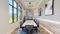 Located at The Reserve in Apopka, Florida, this thoughtfully designed conference room offers a polished and professional environment ideal for meetings and collaborative sessions. The room centers around a long, light wood boardroom table with built-in cable management, surrounded by ergonomic mesh task chairs that comfortably accommodate a large group of participants.

The space is distinguished by its striking overhead lighting — a cascading series of rectangular geometric pendant fixtures in a warm bronze finish — that draw the eye upward and lend a contemporary, upscale character to the room. Flanking the walls, sleek vertical sconce lights add layered illumination and visual depth.

Floor-to-ceiling black-framed windows line one wall, flooding the room with abundant natural light and providing unobstructed views of the surrounding greenery. The interior design blends a neutral palette of whites and soft grays with warm wood-toned wall tile accents running along the lower half of the walls, complemented by a richly patterned area rug underfoot.

Curated artwork and decorative sculptures enhance the room's refined aesthetic, while potted ornamental grasses add organic texture. A glass-paneled sliding door connects the conference room to adjacent common areas, maintaining visual openness. The overall ambiance is modern, well-appointed, and conducive to focused, professional collaboration.