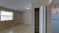 This single-floor space presents a clean and functional interior with a neutral color palette throughout. The room is finished with light beige walls that complement the large-format ceramic tile flooring, lending a bright and cohesive look to the space. A textured popcorn ceiling adds a subtle dimensional quality overhead, with a flush-mount ceiling light fixture providing straightforward illumination. A notable feature is the built-in closet positioned along one wall, fitted with multiple shelves spanning from top to bottom, offering generous and well-organized storage capacity. The bi-fold door design allows for easy access and full visibility of the shelving interior. White baseboards run along the perimeter, providing a clean transition between the walls and the tiled floor. A standard interior door with a classic panel design is visible in the background, along with what appears to be a window, which may allow natural light to enter the space. An electrical outlet is also visible along one wall, indicating practical utility placement. Overall, the space maintains a simple, functional ambiance suited to a variety of uses.