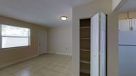 This single-floor space presents a clean and functional interior with a neutral color palette throughout. The room is finished with light beige walls that complement the large-format ceramic tile flooring, lending a bright and cohesive look to the space. A textured popcorn ceiling adds a subtle dimensional quality overhead, with a flush-mount ceiling light fixture providing straightforward illumination. A notable feature is the built-in closet positioned along one wall, fitted with multiple shelves spanning from top to bottom, offering generous and well-organized storage capacity. The bi-fold door design allows for easy access and full visibility of the shelving interior. White baseboards run along the perimeter, providing a clean transition between the walls and the tiled floor. A standard interior door with a classic panel design is visible in the background, along with what appears to be a window, which may allow natural light to enter the space. An electrical outlet is also visible along one wall, indicating practical utility placement. Overall, the space maintains a simple, functional ambiance suited to a variety of uses.